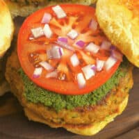 Served burger sized in a soft bun smeared with green chutney, topped with a slice of tomato and chopped red onion.