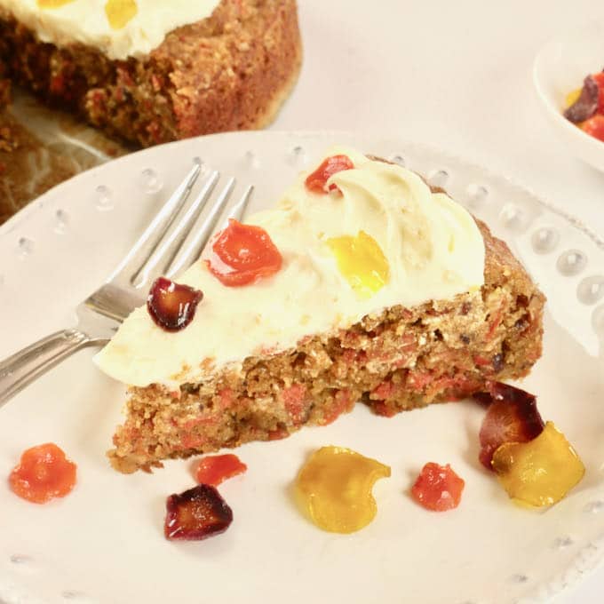 A slice of cake topped with cream cheese frosting and littered with tri-colored candied carrot coins.