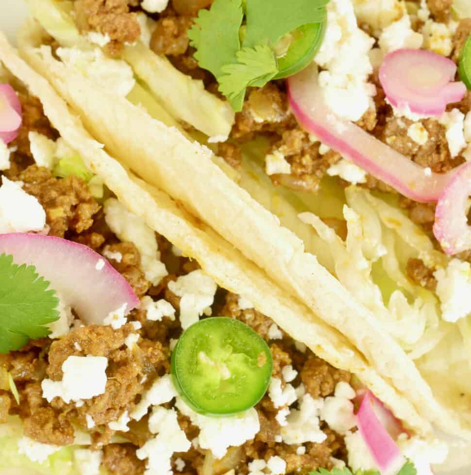 Two tacos garnished pickled red onions, sliced radishes, cheese crumbles and a flutter of cilantro leaves.